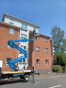 A man works on a lift truck, repairing a roof without scaffolding in front of a building.