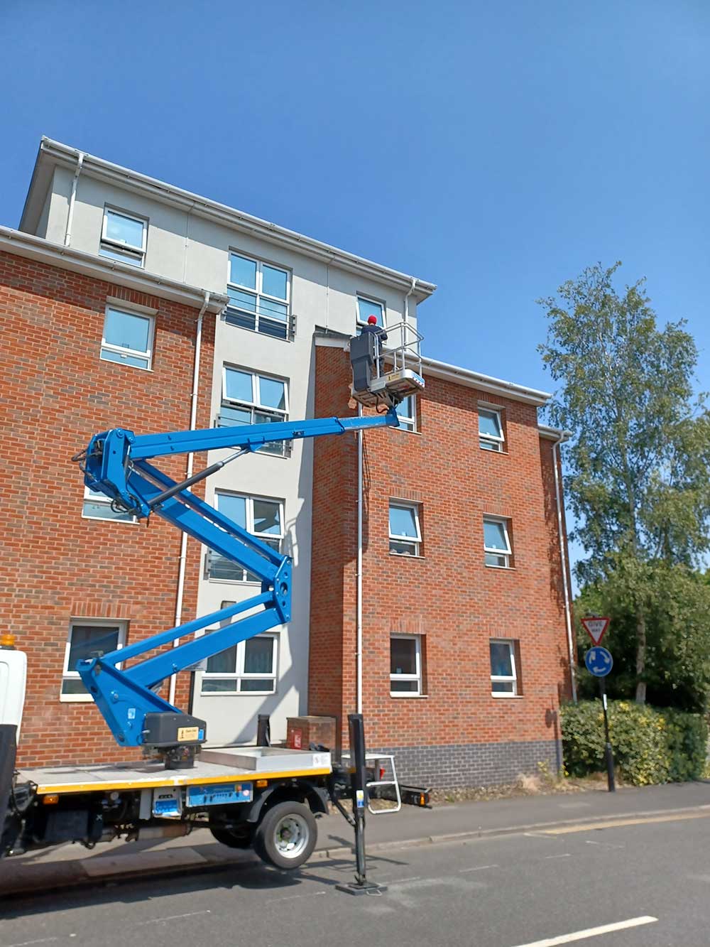 A man works on a lift truck, repairing a roof without scaffolding in front of a building.